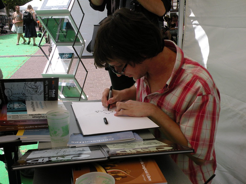 Photo de moi en train de dédicace un tome de GKND sur le stand Framasoft des RMLL 2011 à Strasbourg